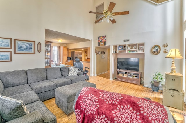 living area featuring a ceiling fan, light wood-style floors, visible vents, and a towering ceiling