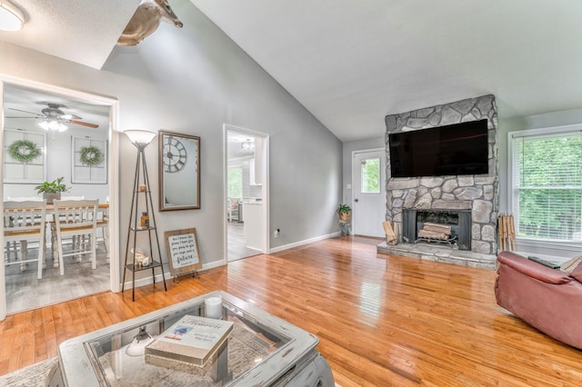 living room featuring a ceiling fan, a stone fireplace, wood finished floors, baseboards, and high vaulted ceiling