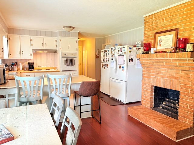 kitchen with white appliances, crown molding, under cabinet range hood, dark wood-style floors, and white cabinetry