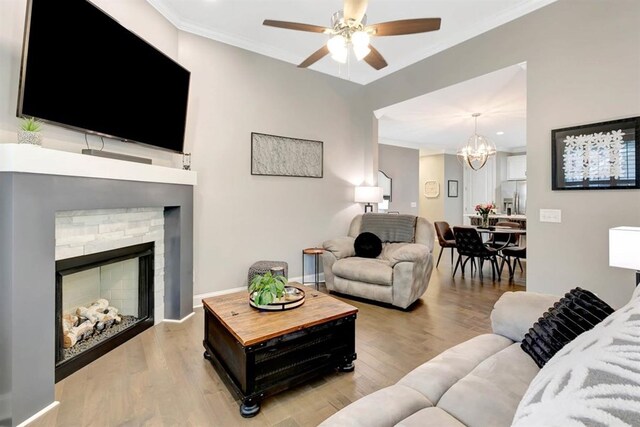 living room featuring crown molding, a stone fireplace, ceiling fan with notable chandelier, and light wood-type flooring