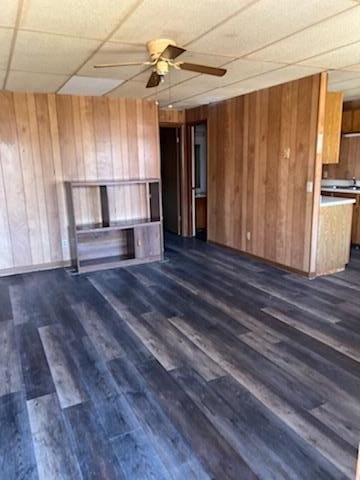 unfurnished living room featuring a drop ceiling, dark hardwood / wood-style floors, ceiling fan, and wood walls