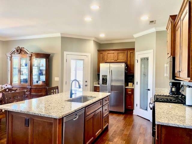 kitchen with light stone counters, dark wood-style flooring, a sink, appliances with stainless steel finishes, and a center island with sink