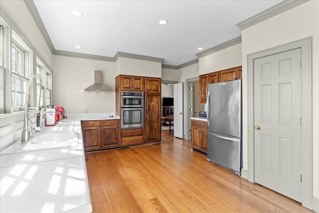 kitchen featuring light wood finished floors, light countertops, appliances with stainless steel finishes, wall chimney range hood, and brown cabinets