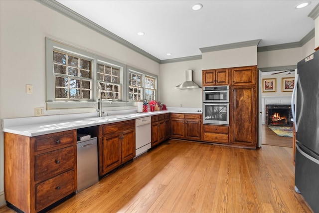 kitchen with a lit fireplace, appliances with stainless steel finishes, light wood-style floors, wall chimney exhaust hood, and a sink