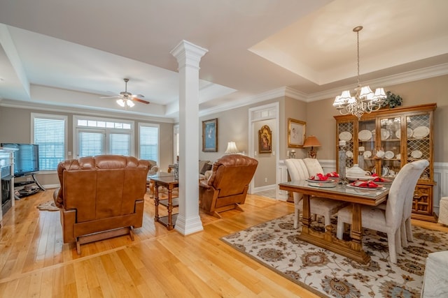 dining space with ceiling fan with notable chandelier, a raised ceiling, ornate columns, and light wood finished floors