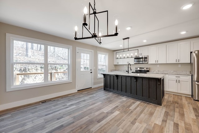 kitchen with appliances with stainless steel finishes, a kitchen island with sink, white cabinets, decorative light fixtures, and light wood-type flooring