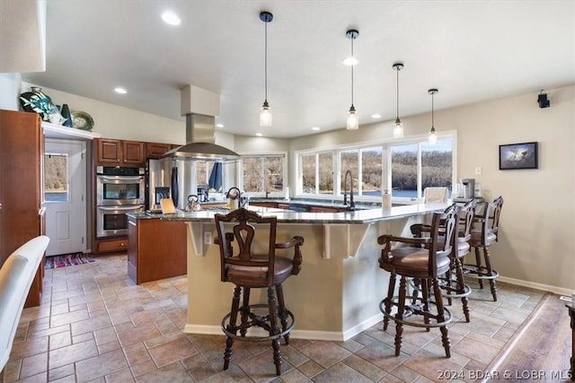 kitchen with a kitchen breakfast bar, island exhaust hood, dark stone countertops, stainless steel double oven, and decorative light fixtures