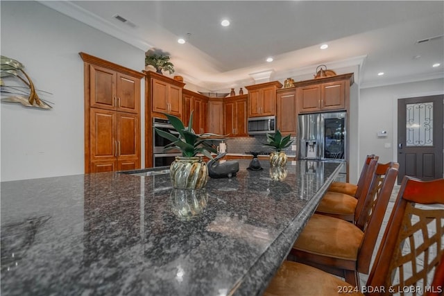 kitchen featuring appliances with stainless steel finishes, tasteful backsplash, dark stone counters, ornamental molding, and a breakfast bar