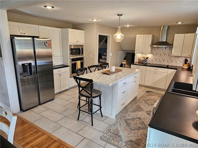 kitchen featuring a center island, decorative light fixtures, stainless steel appliances, wall chimney range hood, and white cabinetry