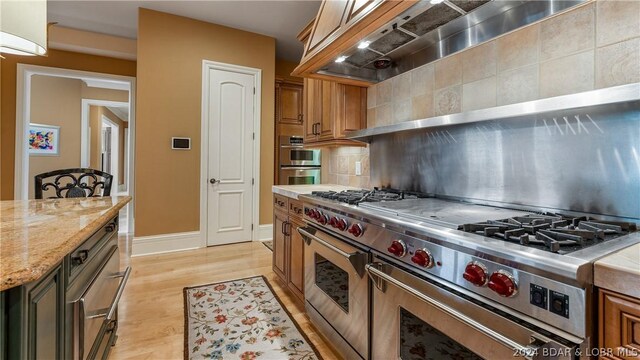 kitchen with light stone countertops, stainless steel appliances, decorative backsplash, premium range hood, and light hardwood / wood-style flooring