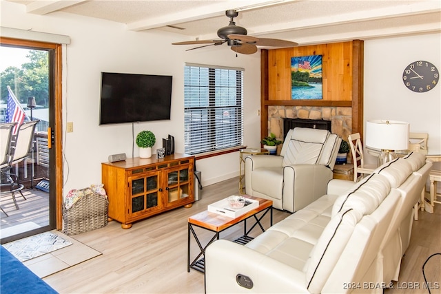 living room with light wood-type flooring, beam ceiling, ceiling fan, and a stone fireplace