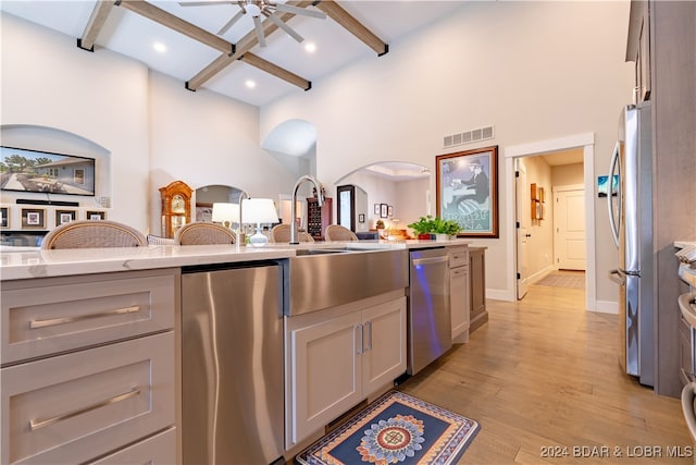 kitchen featuring light wood-type flooring, light stone counters, ceiling fan, appliances with stainless steel finishes, and beam ceiling