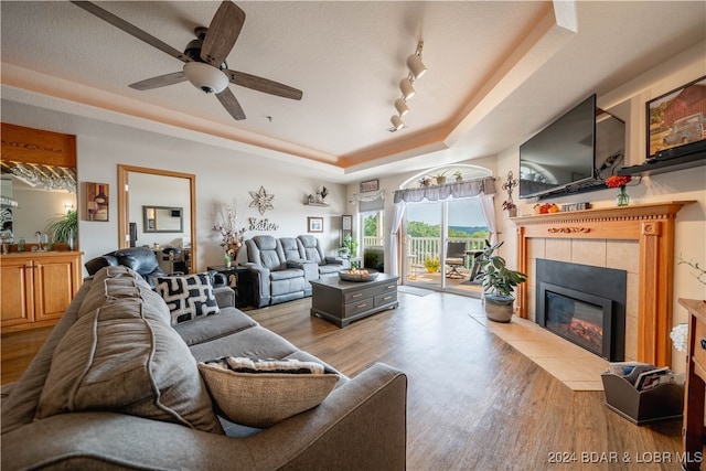 living room featuring a tray ceiling, wood-type flooring, a tile fireplace, and ceiling fan