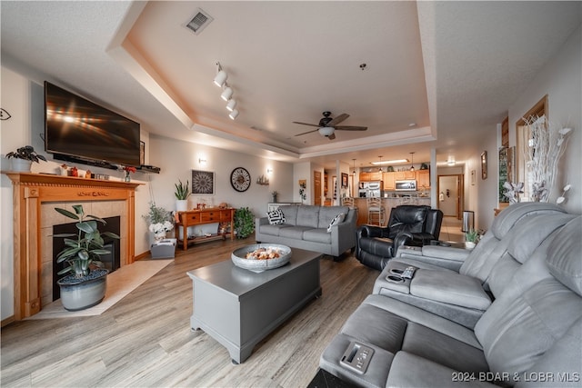 living room with track lighting, a tiled fireplace, ceiling fan, a tray ceiling, and light hardwood / wood-style floors