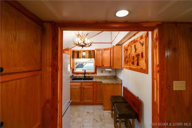 kitchen with light stone countertops, sink, white refrigerator, a notable chandelier, and wood walls