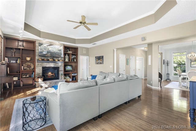 living room with dark wood-type flooring, ceiling fan with notable chandelier, a stone fireplace, a raised ceiling, and ornamental molding