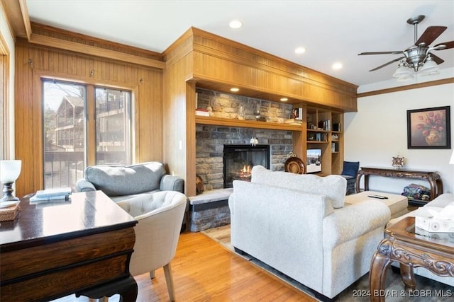 living room featuring wood walls, a stone fireplace, crown molding, ceiling fan, and light wood-type flooring