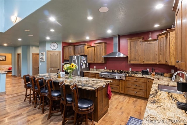 kitchen featuring an island with sink, sink, light hardwood / wood-style flooring, wall chimney range hood, and appliances with stainless steel finishes