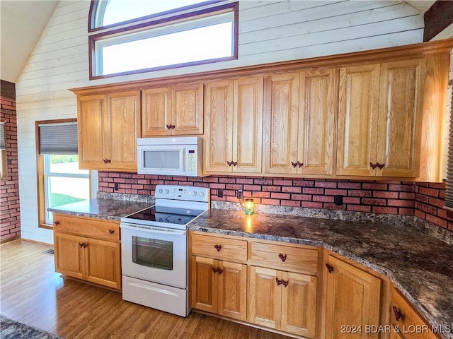 kitchen with wood walls, white appliances, brick wall, light hardwood / wood-style flooring, and dark stone counters