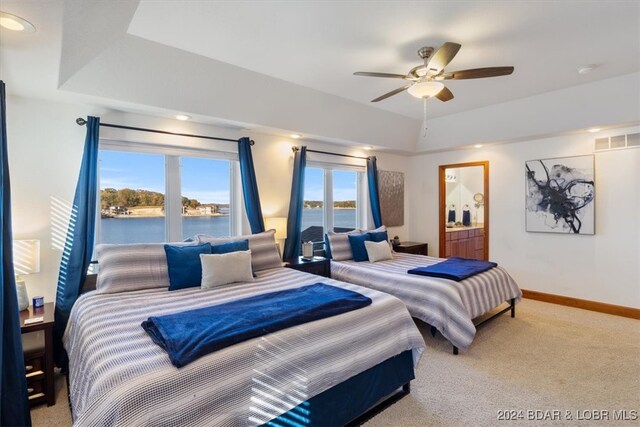 carpeted bedroom featuring a raised ceiling, ensuite bathroom, a water view, and ceiling fan