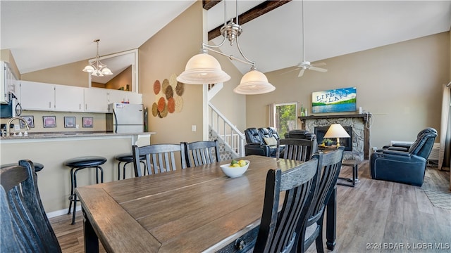 dining area featuring beam ceiling, high vaulted ceiling, light wood-type flooring, a fireplace, and ceiling fan with notable chandelier