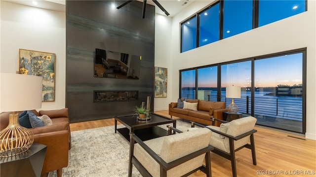 living room featuring ceiling fan with notable chandelier, a water view, hardwood / wood-style floors, a large fireplace, and a high ceiling