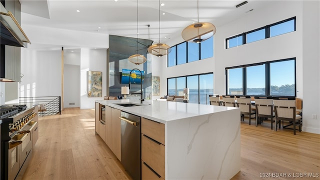 kitchen featuring stainless steel appliances, light wood-type flooring, pendant lighting, light stone countertops, and an island with sink