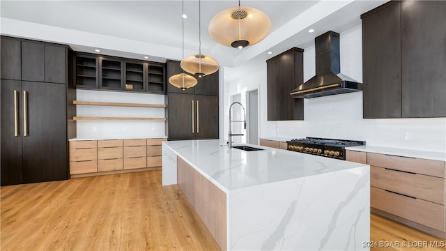 kitchen with light stone counters, wall chimney exhaust hood, decorative light fixtures, an island with sink, and stove