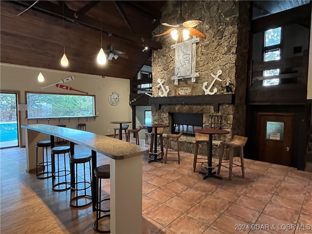 kitchen featuring ceiling fan, a breakfast bar area, high vaulted ceiling, a stone fireplace, and pendant lighting