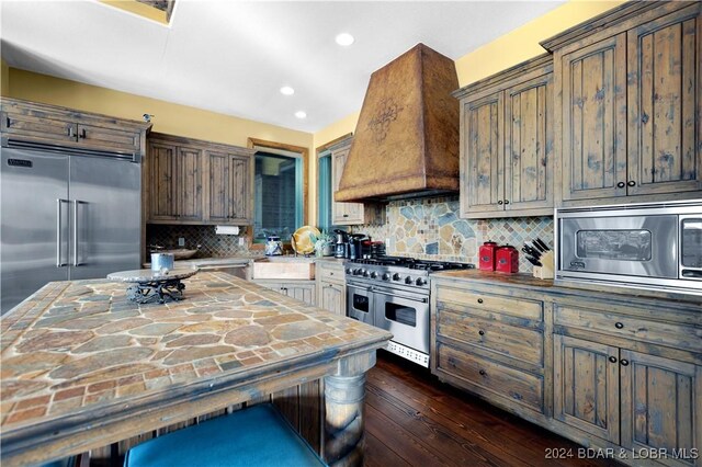 kitchen with built in appliances, backsplash, dark wood-type flooring, and custom exhaust hood