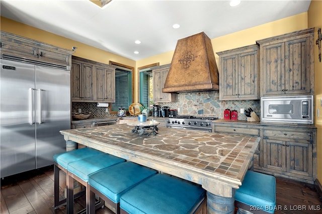 kitchen with custom exhaust hood, dark wood-type flooring, backsplash, built in appliances, and a kitchen island