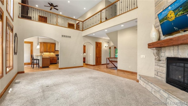 unfurnished living room with sink, a stone fireplace, a towering ceiling, ceiling fan, and light carpet