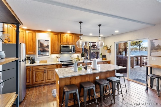 kitchen with light stone countertops, hanging light fixtures, dark hardwood / wood-style floors, a center island with sink, and appliances with stainless steel finishes