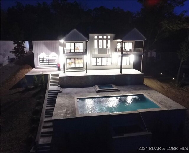 back house at night with a swimming pool with hot tub, a patio, and a balcony