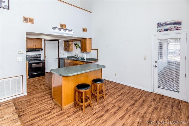 kitchen with a breakfast bar, black electric range oven, stainless steel dishwasher, hardwood / wood-style flooring, and a towering ceiling