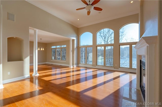 unfurnished living room featuring a high end fireplace, decorative columns, ceiling fan with notable chandelier, crown molding, and hardwood / wood-style floors