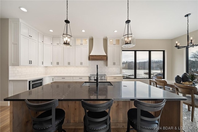 kitchen with custom exhaust hood, a kitchen island with sink, sink, wood-type flooring, and white cabinetry