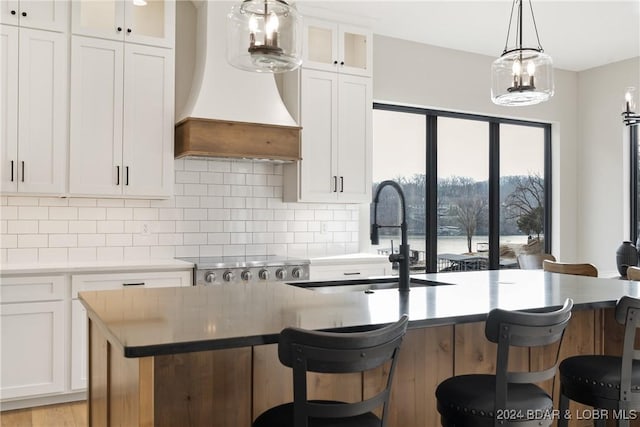 kitchen with pendant lighting, custom exhaust hood, a kitchen island with sink, white cabinets, and decorative backsplash