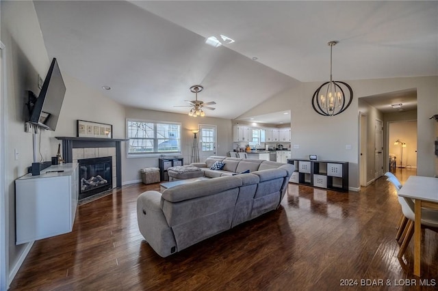 living room with ceiling fan with notable chandelier, dark hardwood / wood-style flooring, a tiled fireplace, and vaulted ceiling