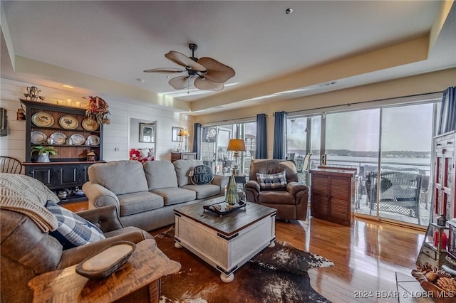 living room with a tray ceiling, wooden walls, ceiling fan, and wood-type flooring