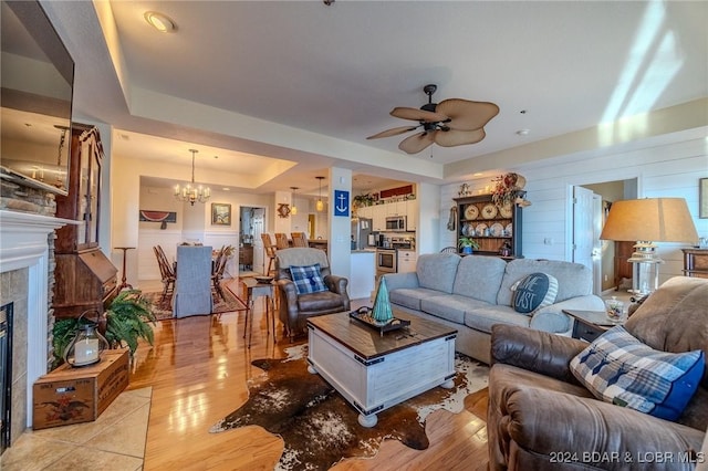 living room featuring wood walls, ceiling fan with notable chandelier, light wood-type flooring, a tray ceiling, and a tiled fireplace