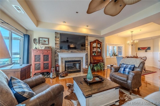 living room featuring a fireplace, light wood-type flooring, a tray ceiling, and ceiling fan with notable chandelier