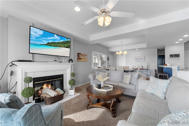 living room with a raised ceiling, a tiled fireplace, light carpet, and ceiling fan with notable chandelier