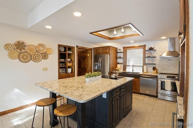 kitchen with a center island with sink, a kitchen breakfast bar, a raised ceiling, wall chimney range hood, and appliances with stainless steel finishes