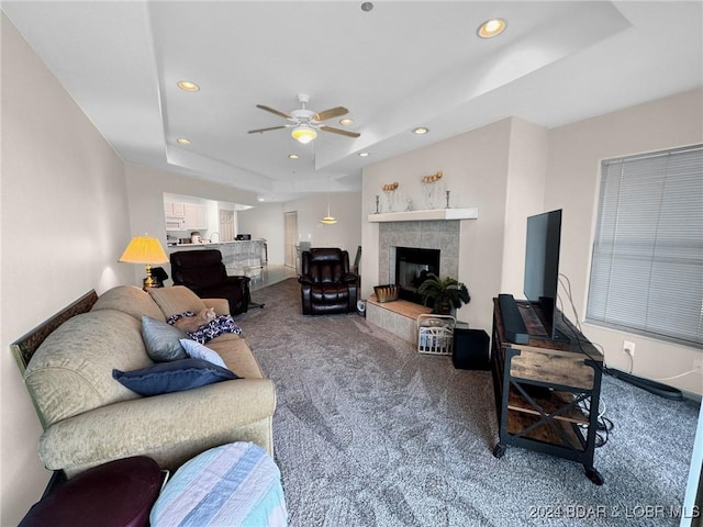 carpeted living room with a raised ceiling, a tile fireplace, and ceiling fan