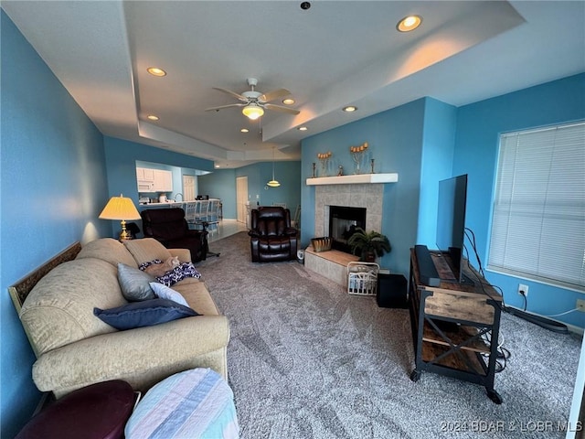 carpeted living room featuring a tray ceiling, a tiled fireplace, and ceiling fan