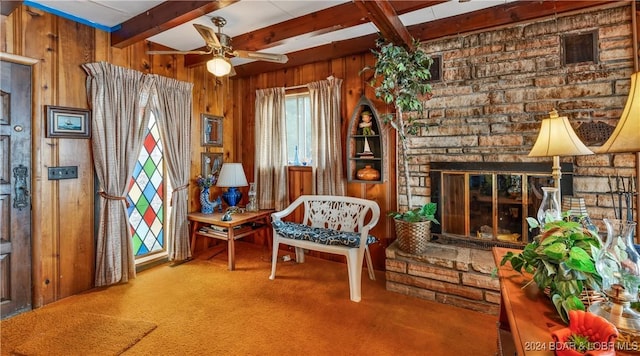 sitting room featuring ceiling fan, wooden walls, beam ceiling, carpet floors, and a stone fireplace