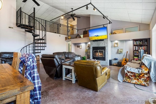 living area featuring finished concrete flooring, a glass covered fireplace, ceiling fan, high vaulted ceiling, and stairs