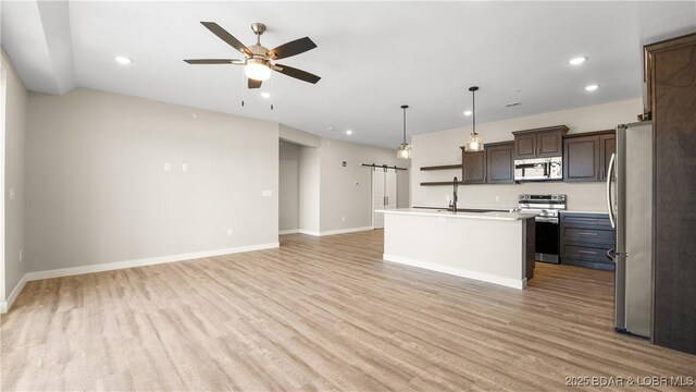 kitchen with appliances with stainless steel finishes, an island with sink, decorative light fixtures, a barn door, and light wood-type flooring