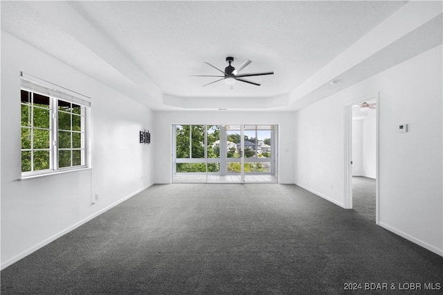 empty room with ceiling fan, a wealth of natural light, a tray ceiling, and dark colored carpet